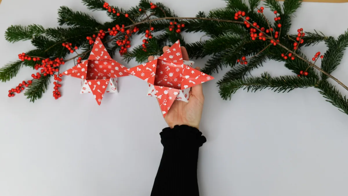 Anleitung Origami Box selber machen. Eine Hand hält eine aus weihnachtlichem Papier gefaltene Box in Sternform. Im Hintergrund grüne Äste mit Nadeln und rote Früchte.