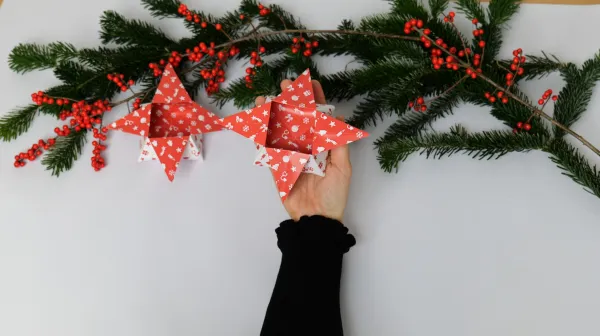 Anleitung Origami Box selber machen. Eine Hand hält eine aus weihnachtlichem Papier gefaltene Box in Sternform. Im Hintergrund grüne Äste mit Nadeln und rote Früchte.